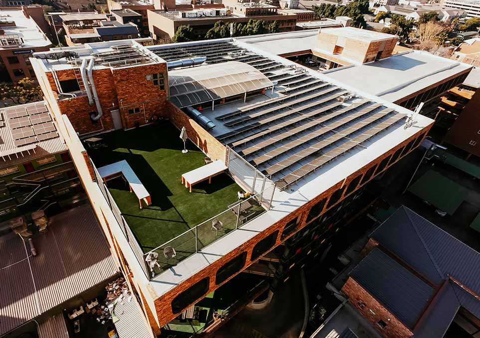 NXT Gen: aerial view of the completed brick commercial building, showing the rooftop solar array and the artificial turf roof terrace.